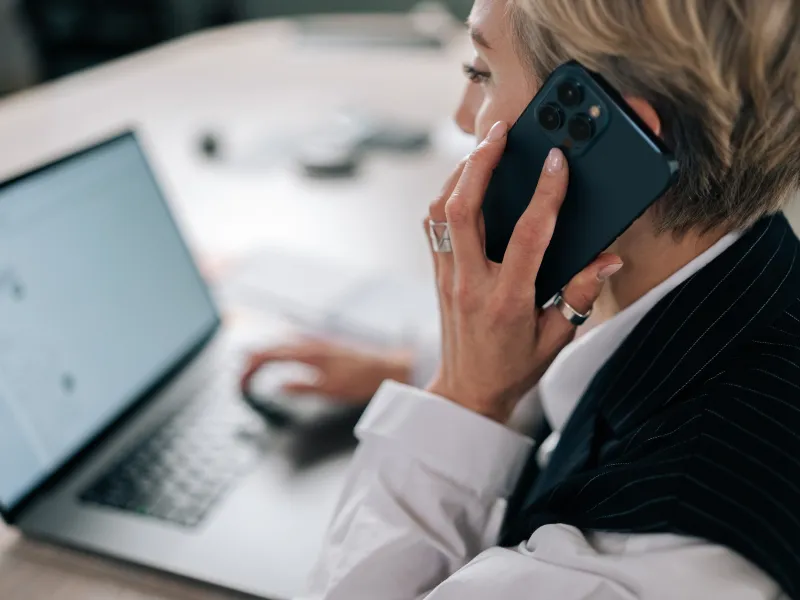 Professional woman in business attire talking on smartphone while working on a laptop at desk.