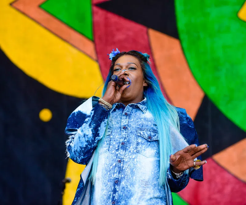 Female singer with blue ombre hair performing on stage with colorful abstract background art.