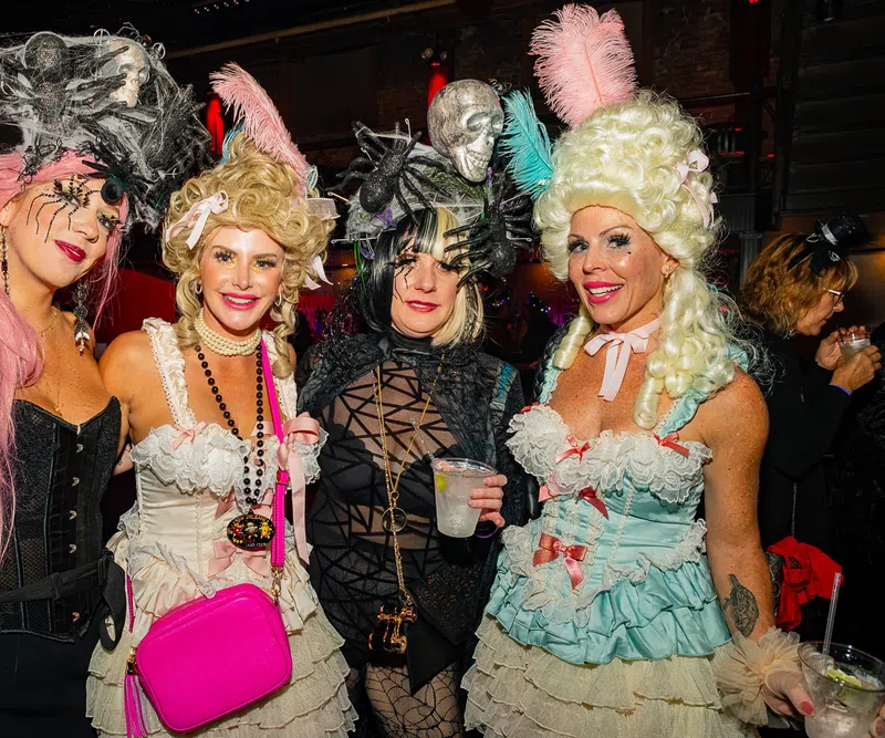 Four women in elaborate Halloween costumes with wigs and accessories at a festive party indoors.