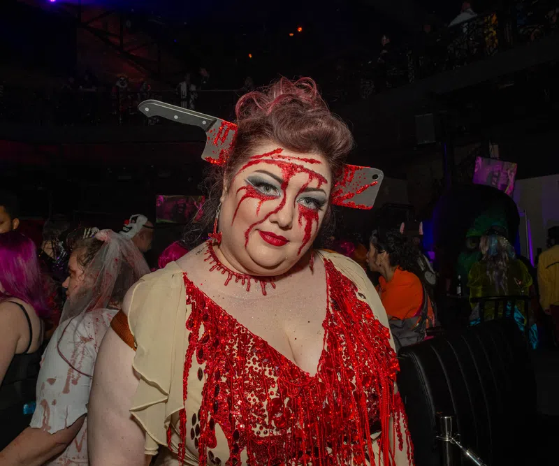 Woman in bloody costume with fake cleaver through head at a Halloween party with crowd in background
