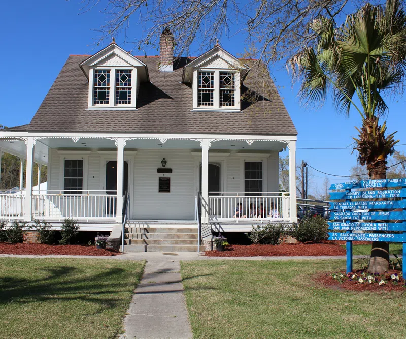 White historic house with a wraparound porch, manicured lawn, and a blue directional sign near a palm tree.