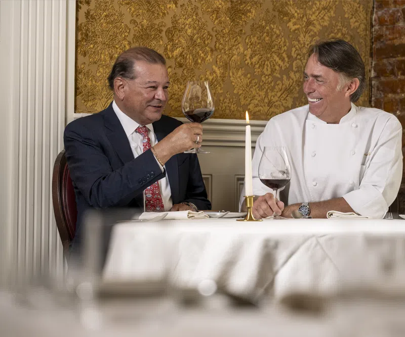 Two men smiling and toasting with red wine in an elegant dining room with gold wallpaper and exposed brick.