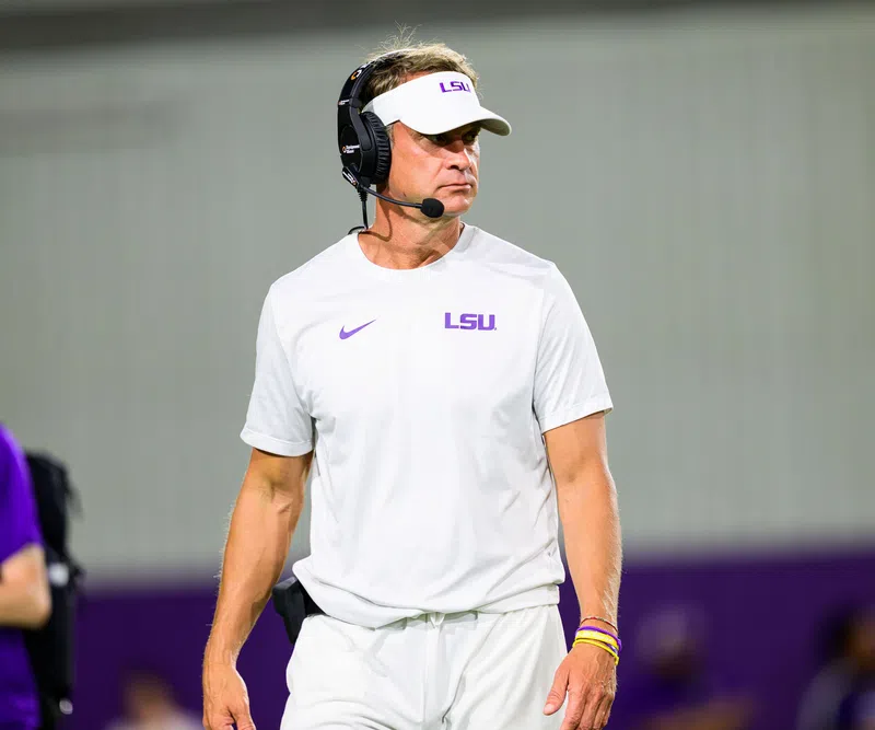 LSU football coach in white LSU gear and visor wears a headset on the sidelines during a game.