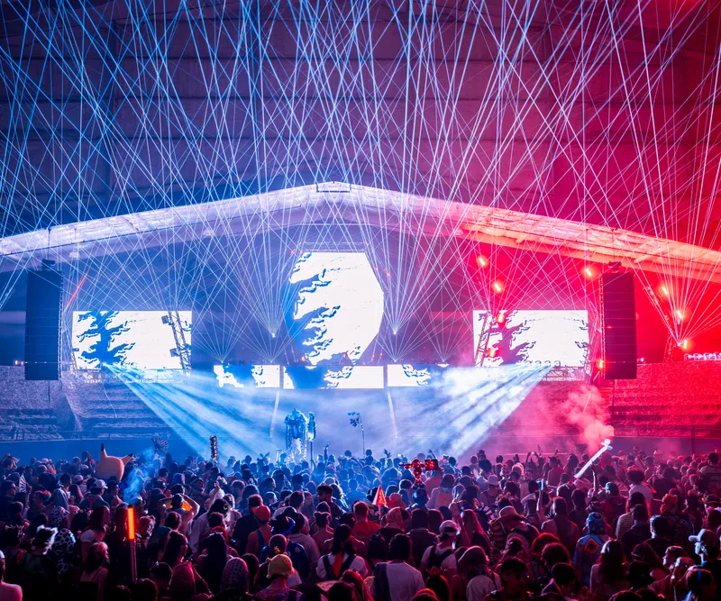 Crowd enjoying a nighttime concert with vibrant laser lights and colorful stage effects under a large canopy.