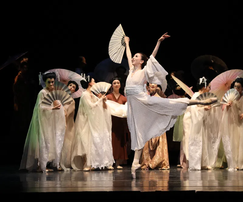 Ballet dancer in white costume performs with fans on stage with other dancers in traditional attire.