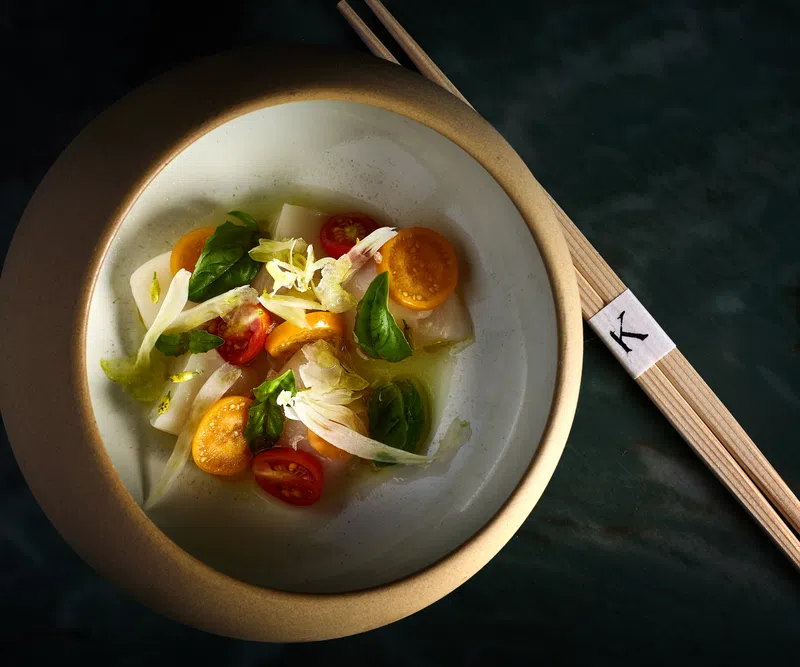 Elegant bowl of fresh vegetable salad with cherry tomatoes, greens, and chopsticks on dark surface.