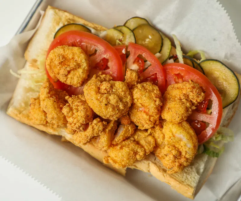 Fried shrimp po'boy sandwich with sliced tomatoes, pickles, and lettuce on a sub roll.