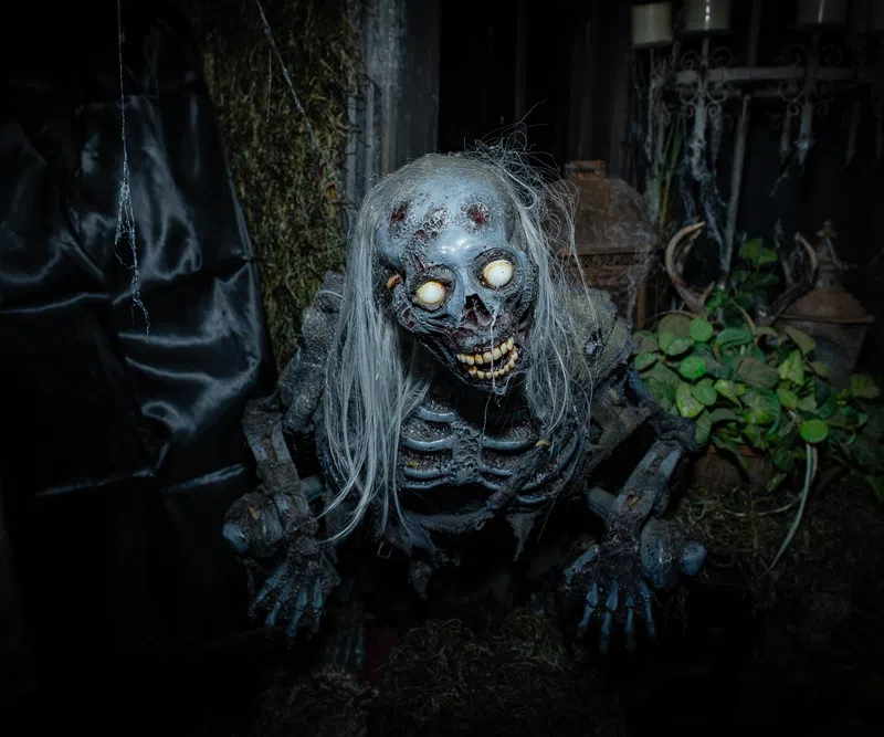 Creepy skeleton figure with glowing eyes and long white hair in a dark, eerie setting with plants and moss.
