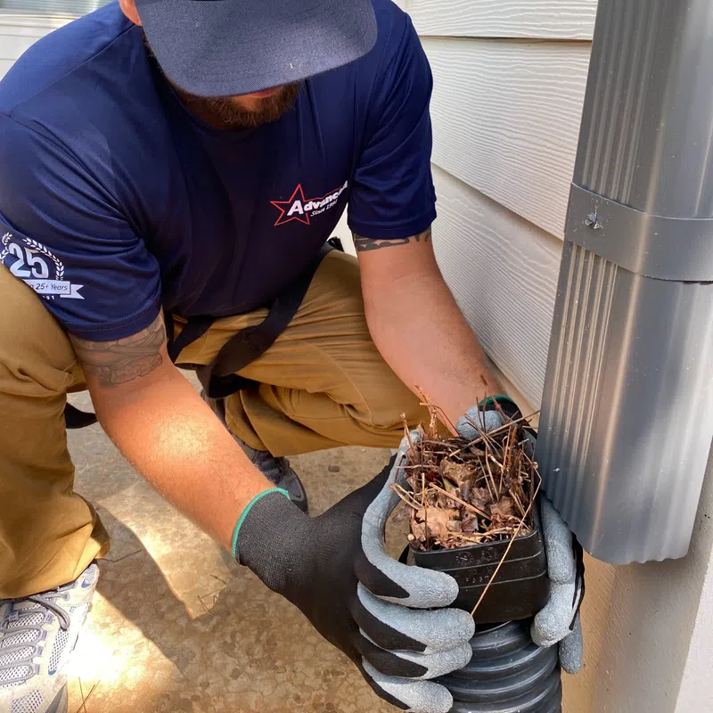 A worker wearing gloves removes debris from a drainage system near a house.