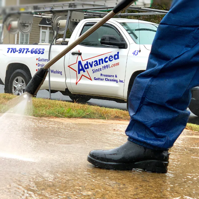 Person using a pressure washer on a driveway with a cleaning service truck in the background.