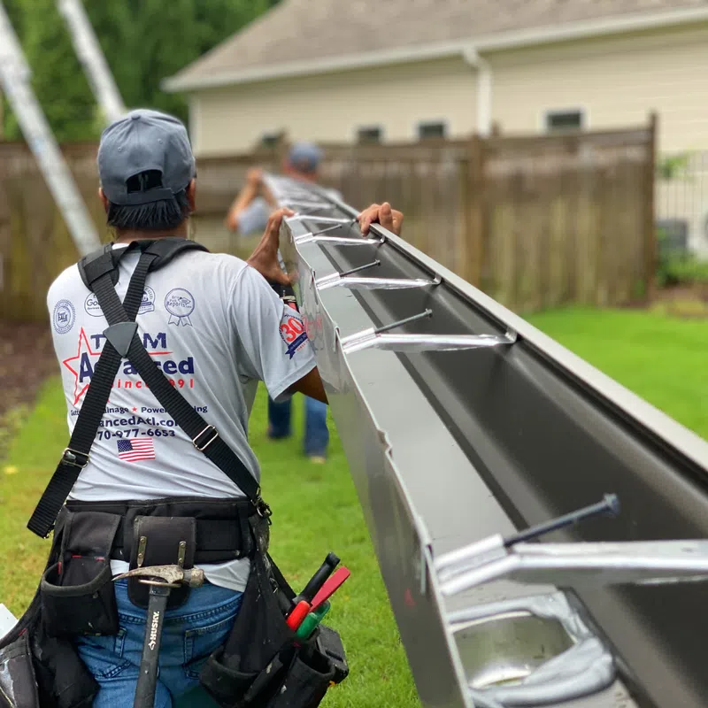 Worker installing gutters on a house with a ladder in the background, showcasing home maintenance.
