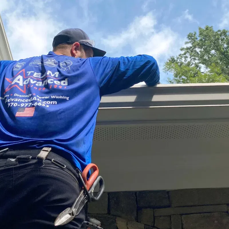 A worker in a blue shirt cleaning gutters under a clear sky with trees in the background.