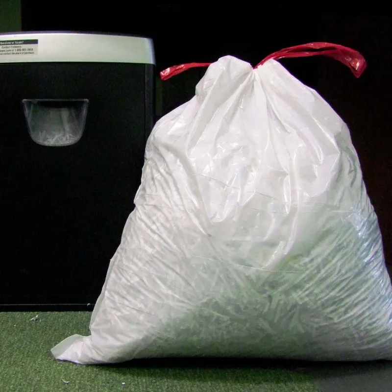 Large white plastic bag full of shredded paper stands next to a black paper shredder on green carpet.