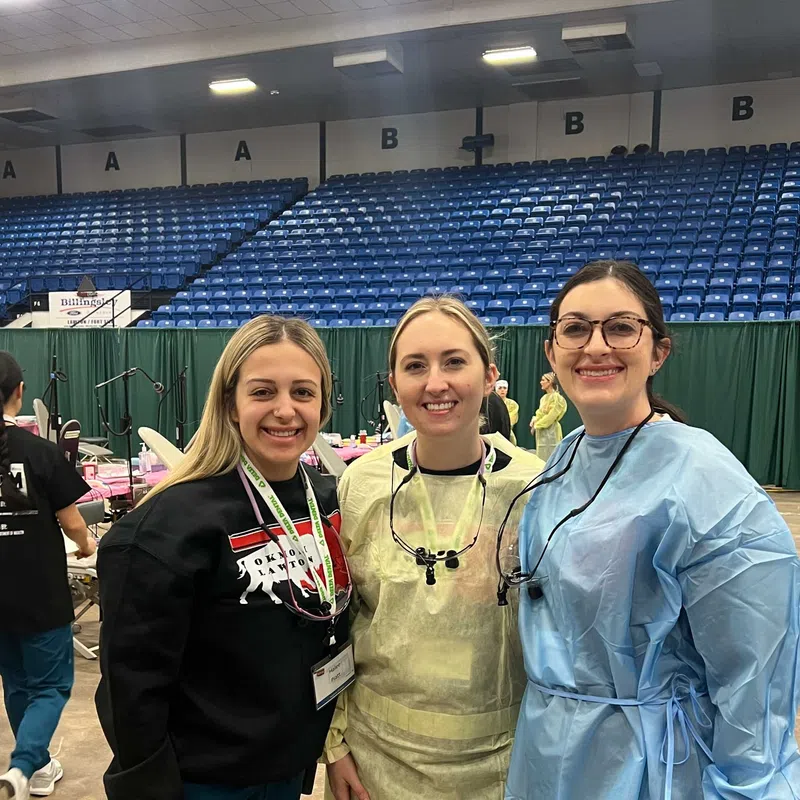 Three women smiling at a medical event, two wearing protective gowns and one in casual clothes inside a large venue.