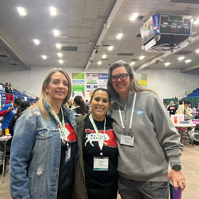 Three women smiling and posing together at an indoor event with name tags and casual clothing under bright lights.