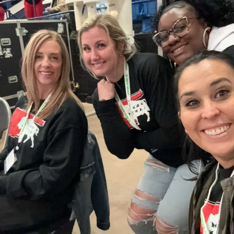 Four women smiling and posing for a selfie in a casual indoor event setting with lanyards and black shirts.