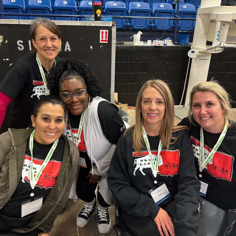 Group of five smiling women wearing event badges and matching Oklahoma Law 2024 shirts at an indoor venue.