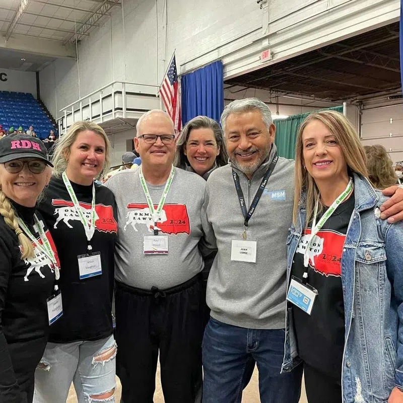 Group of six smiling adults wearing event badges pose together in an indoor arena with blue seating and USA flag.