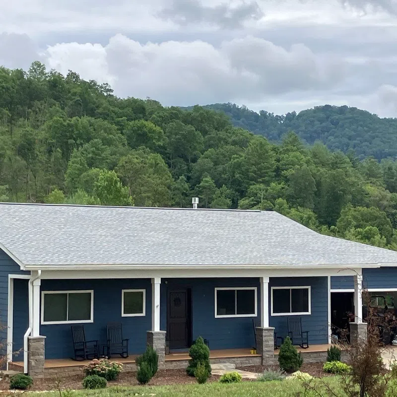 Blue ranch-style house with porch and rocking chairs set against green forested hills and cloudy sky.