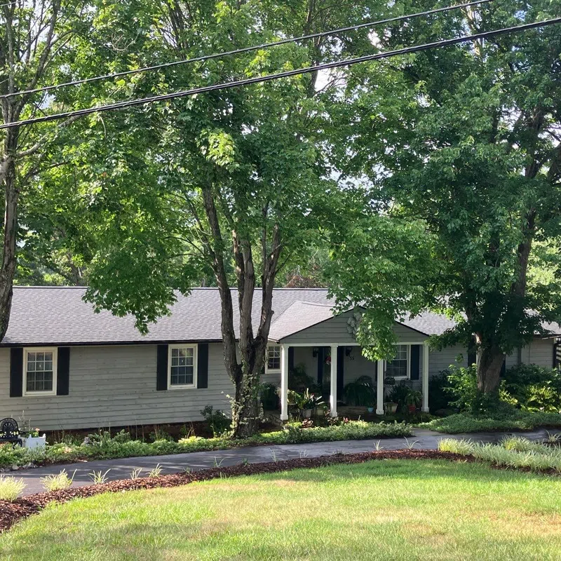 Single-story gray house with black shutters surrounded by green trees and a well-kept lawn on a sunny day.