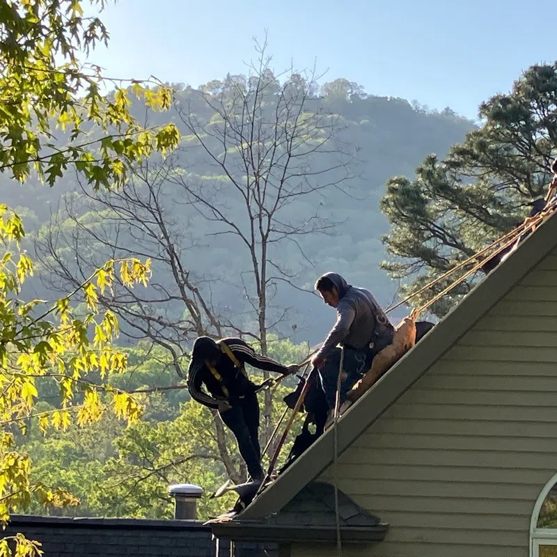 Workers secured with ropes repair the steep roof of a house surrounded by trees and hills under a clear sky.