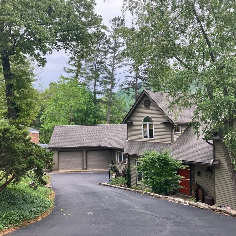 Two-story house with gray siding, red front door, double garage, surrounded by lush green trees and a paved driveway.