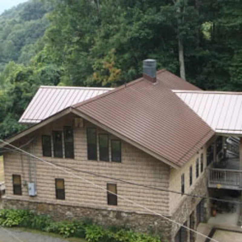 Rustic house with brown metal roof and stone foundation surrounded by lush green forest on a hillside.