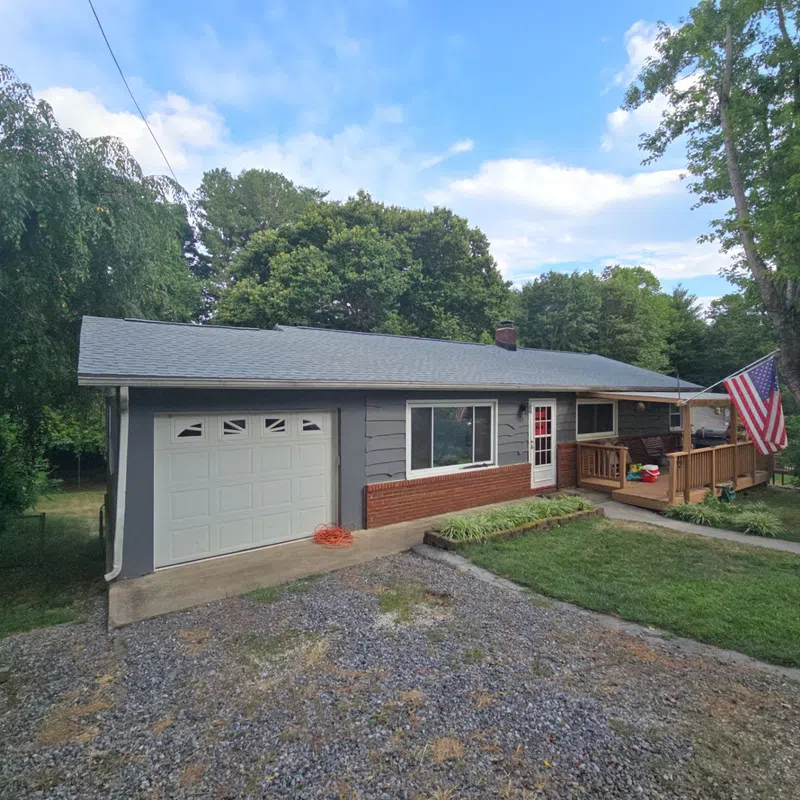 Single-story house with gray siding, brick accent, front porch with American flag, and gravel driveway surrounded by trees.