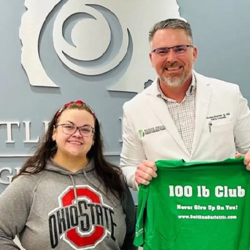 Smiling woman in Ohio State hoodie stands next to man in white coat holding green 100 lb Club shirt