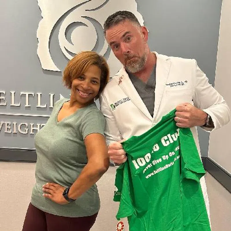 Smiling woman posing with a doctor holding a green shirt in a weight loss clinic reception area.