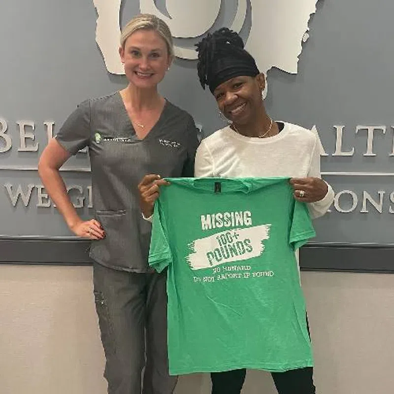 Two smiling women stand indoors, one in gray scrubs, the other holding a green shirt reading Missing 100+ pounds.