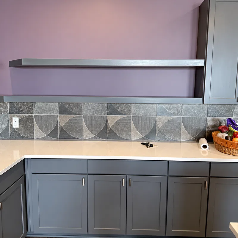 Modern kitchen corner with purple walls, gray cabinetry, geometric tiled backsplash, and a fruit basket on countertop