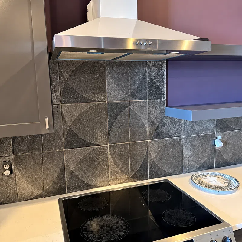 Modern kitchen stove with black glass cooktop, stainless steel range hood, and geometric patterned tile backsplash.