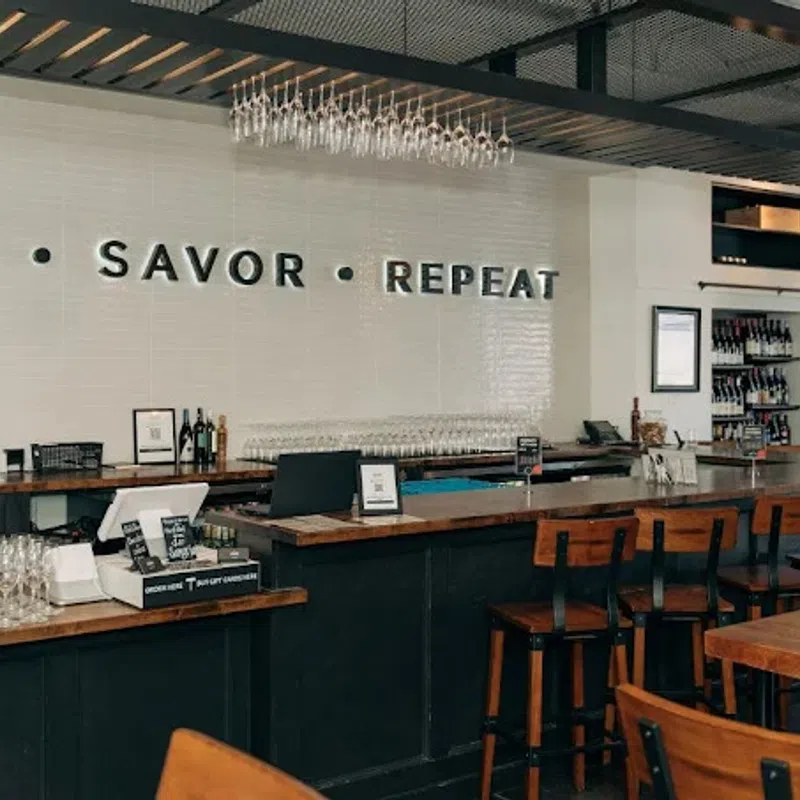 Modern bar interior with wooden stools, wine glasses, and wall text reading SIP SAVOR REPEAT above counter.