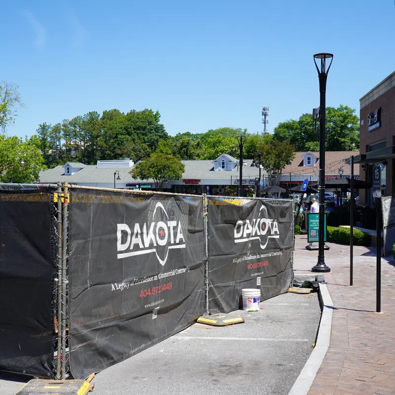 Construction site fenced with black Dakota-branded barriers in a sunny outdoor shopping area with trees and buildings.