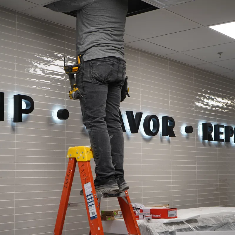 Worker on a ladder installing illuminated wall letters reading SIP VOR REPEAT on tiled wall.