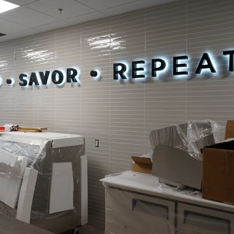 Illuminated SIP SAVOR REPEAT sign on tiled wall in room with covered furniture and unpacked boxes.