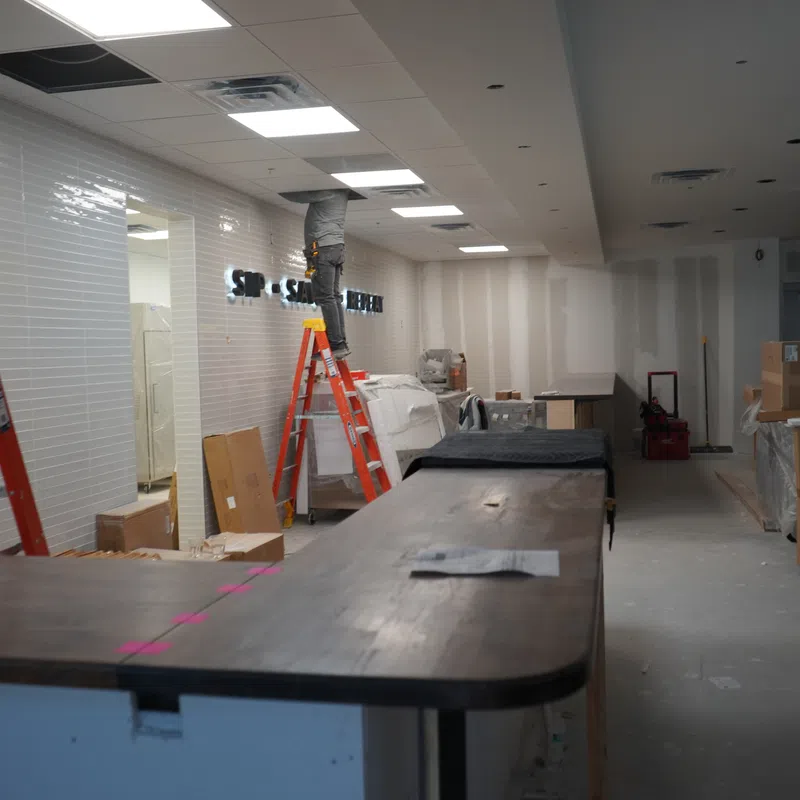 Under-construction interior with ladders, tools, boxes, and a worker installing fixtures in a commercial space.
