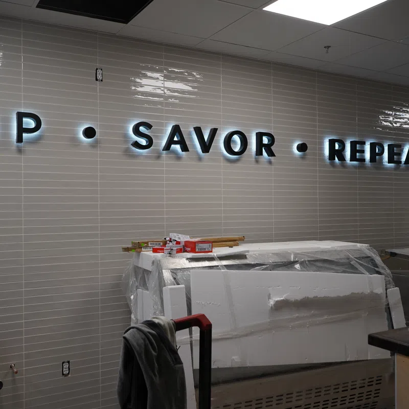 Illuminated wall sign reading SIP • SAVOR • REPEAT over a kitchen countertop with covered appliances and prep area.