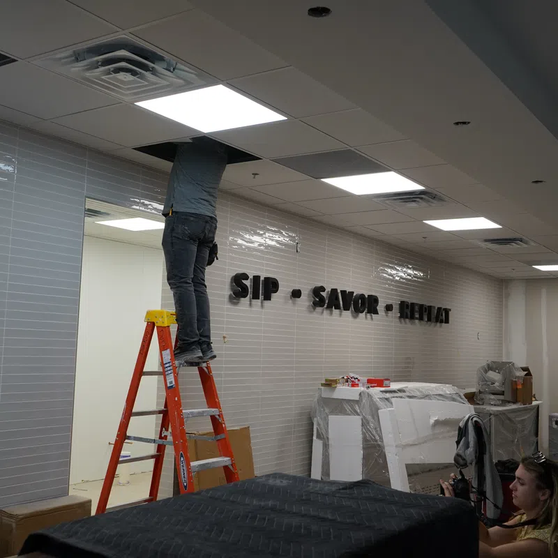 Worker on ladder installing ceiling tiles in a room with tiled walls displaying 'SIP SAVOR RELAX' sign.