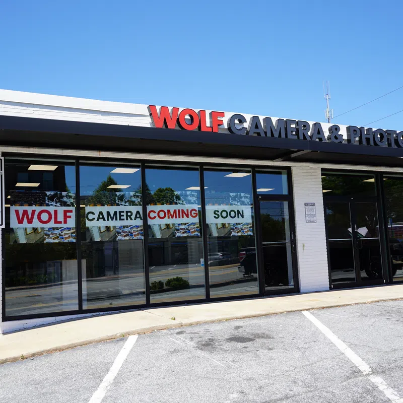 Storefront of Wolf Camera & Photo with signs announcing new store coming soon under clear blue sky.