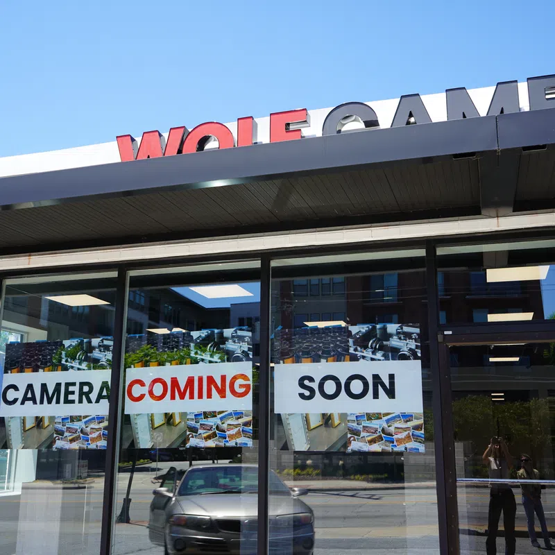 Storefront with signs in windows announcing Wolf Camera coming soon under a clear blue sky.