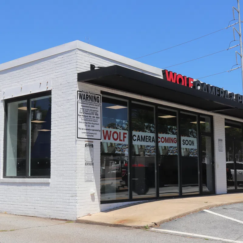 White brick storefront with glass windows displaying Wolf Camera coming soon signs under clear blue sky