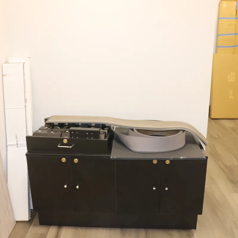 Black printer or copier machine in a room with wooden floor and white walls, surrounded by stacked boxes and panels