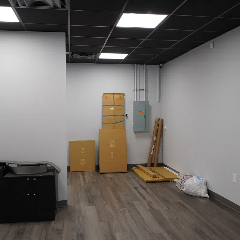Empty room with light wood flooring, gray walls, black ceiling tiles, cardboard boxes, and paint supplies on the floor.