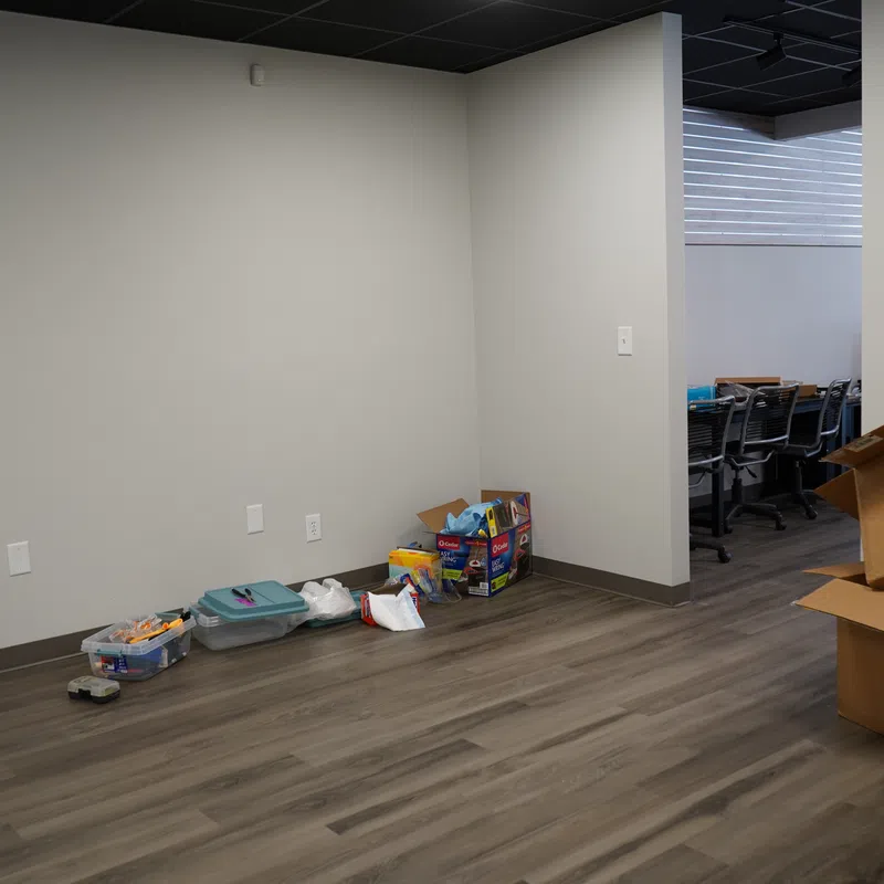 Empty office corner with unpacked boxes, paint buckets, and plastic containers on wooden floor near white walls.