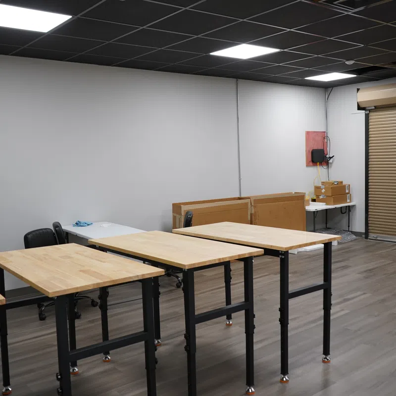 Empty workshop room with wooden tables, black chairs, gray walls, and a roll-up door on wood flooring.