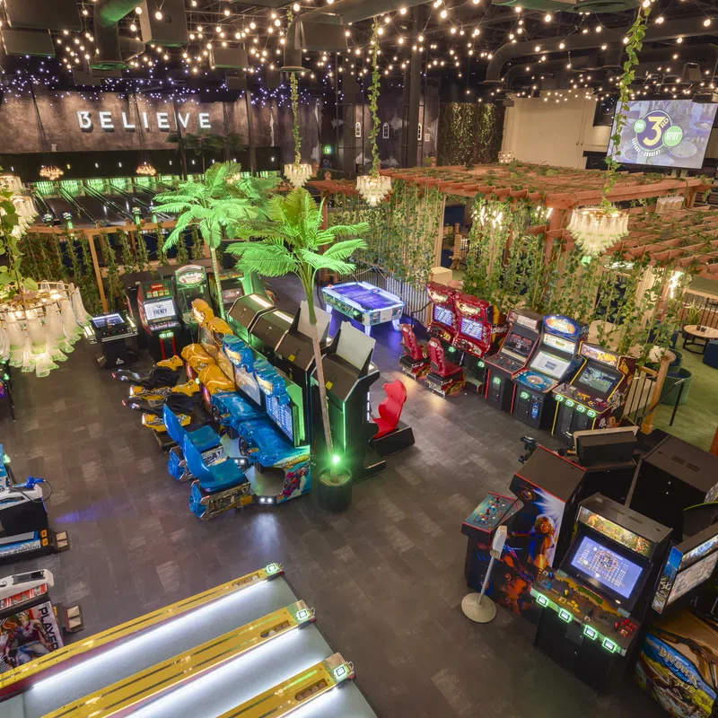 Indoor arcade with vintage and modern game machines, colorful seats, green plants, and string lights above.