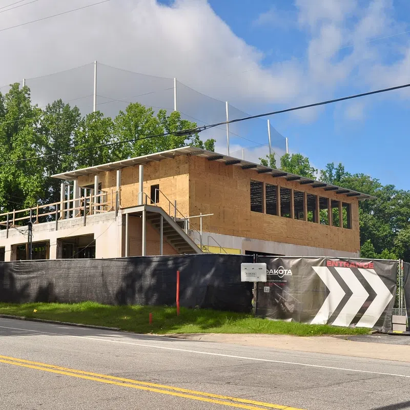 Two-story building under construction with plywood walls and black fence, surrounded by trees and clear sky.