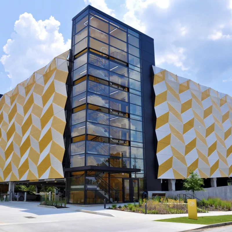 Modern parking garage with geometric yellow and white facade and central glass staircase under a blue sky with clouds.